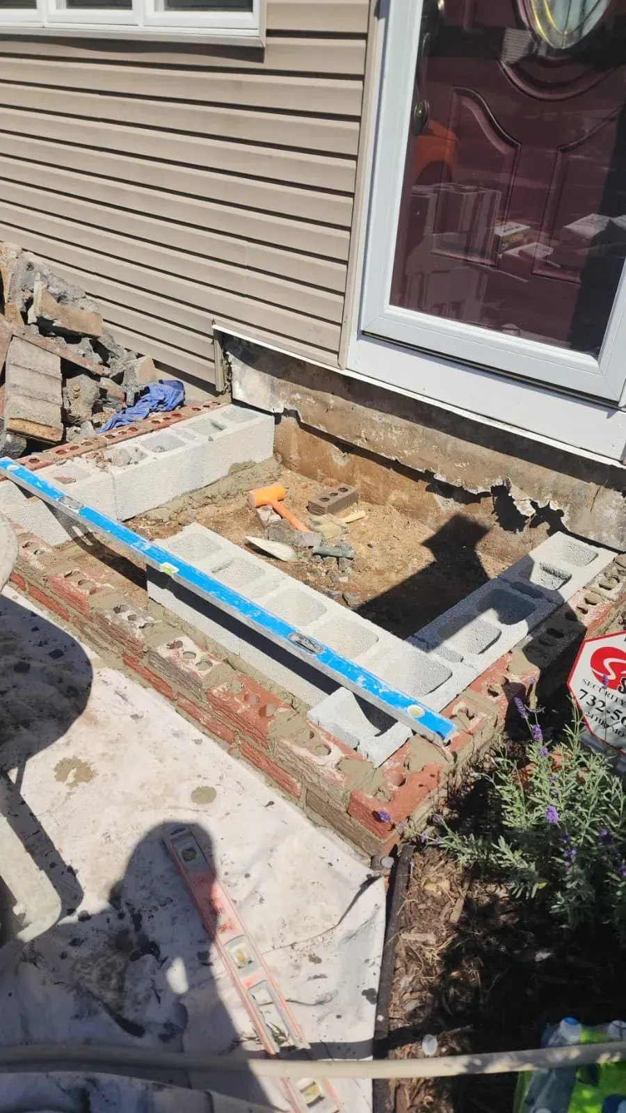 Partially demolished concrete porch beside tan siding and a glass door, with exposed foundation and blue framing boards