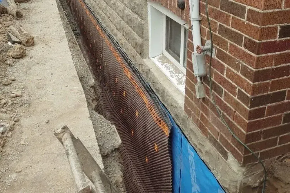 Brick house foundation with black mesh and blue tarp covering an excavated trench along the wall