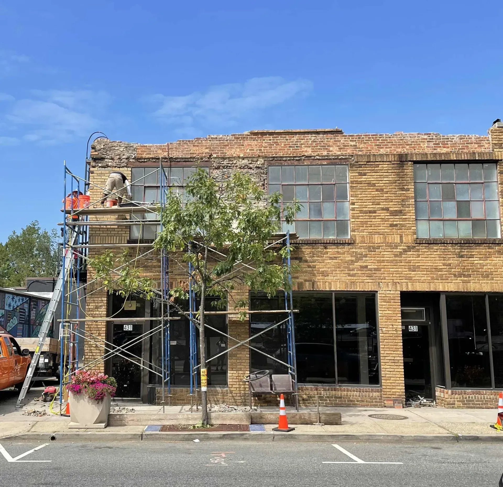 Scaffolding surrounds a brick storefront under renovation on a sunny street, with trees and an orange cone nearby.