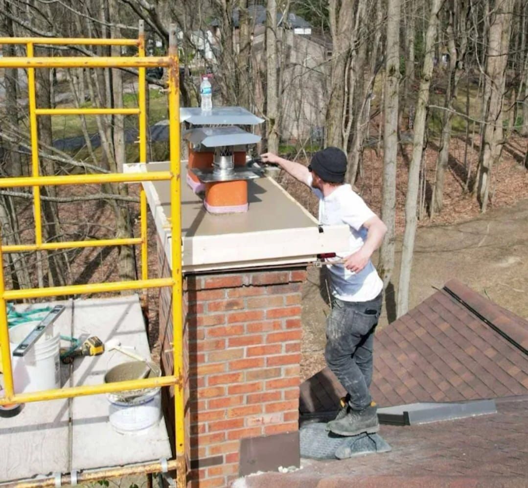 Man on a roof works on a chimney with a scaffold. He's wearing a hat, checking a paper. Sunny day, trees in background.