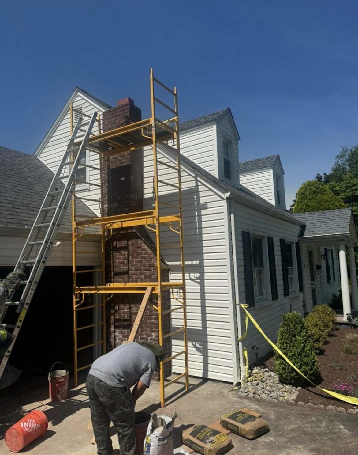A person repairing a brick chimney on a white house, with scaffolding, ladder, and supplies on a sunny day.