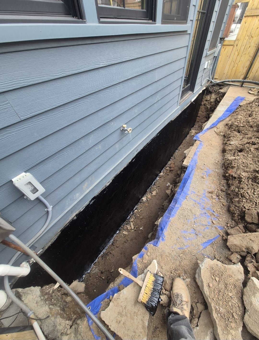 A trench dug along a building's blue siding, with black sealant visible. A brush sits nearby.