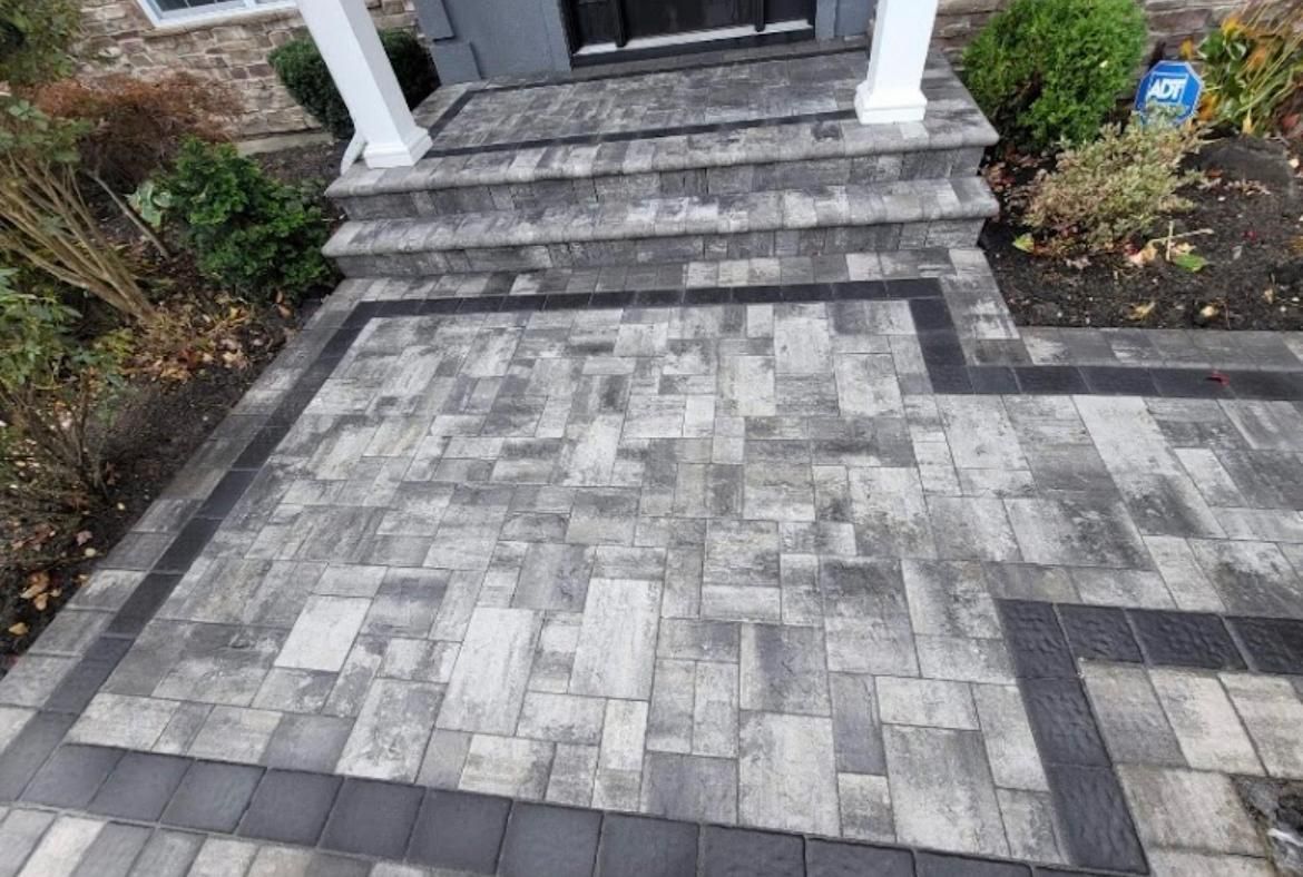 Brick walkway leading to home's steps; gray bricks with black border, small plants and grass on sides.