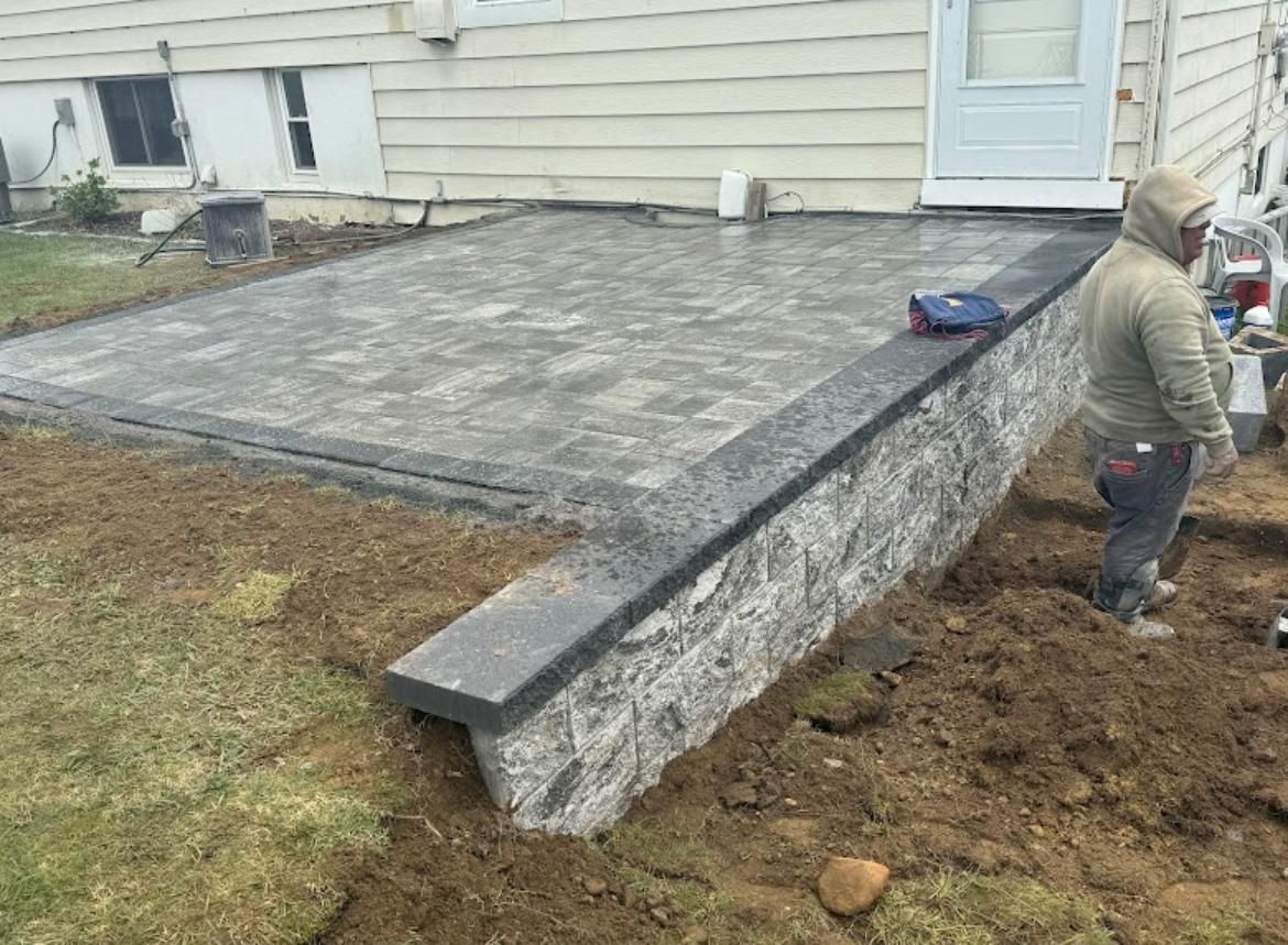 Man builds a stone patio next to a house. A low stone wall and grey pavers are visible.