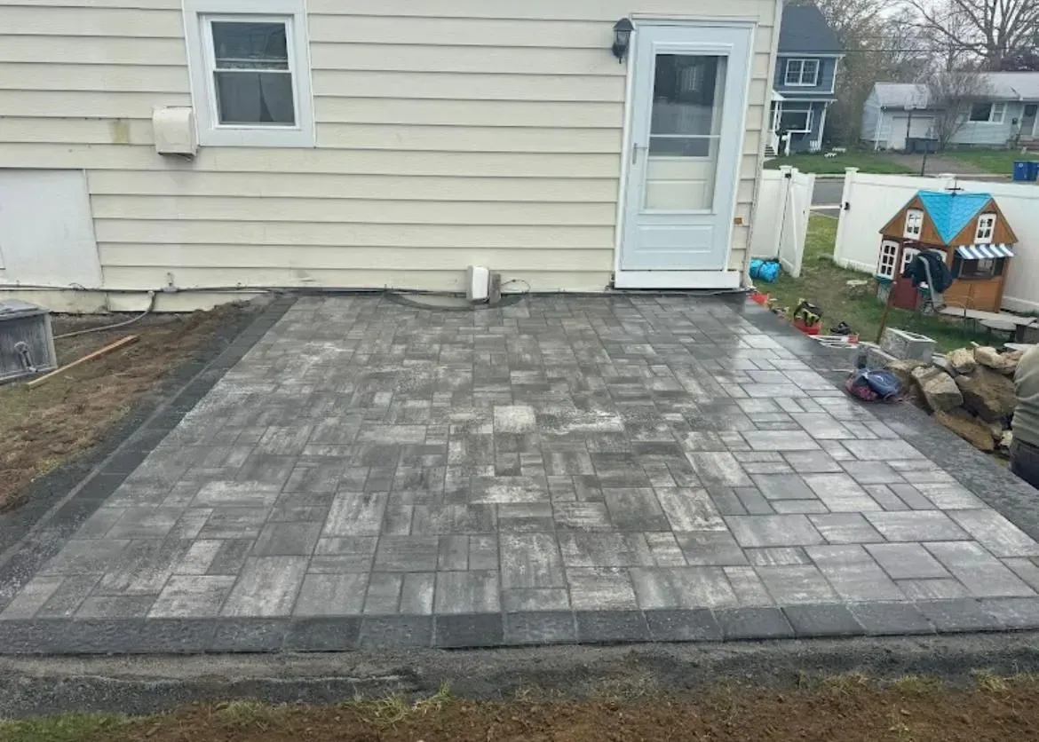 Gray paver patio beside a house with a white door and small garden area.