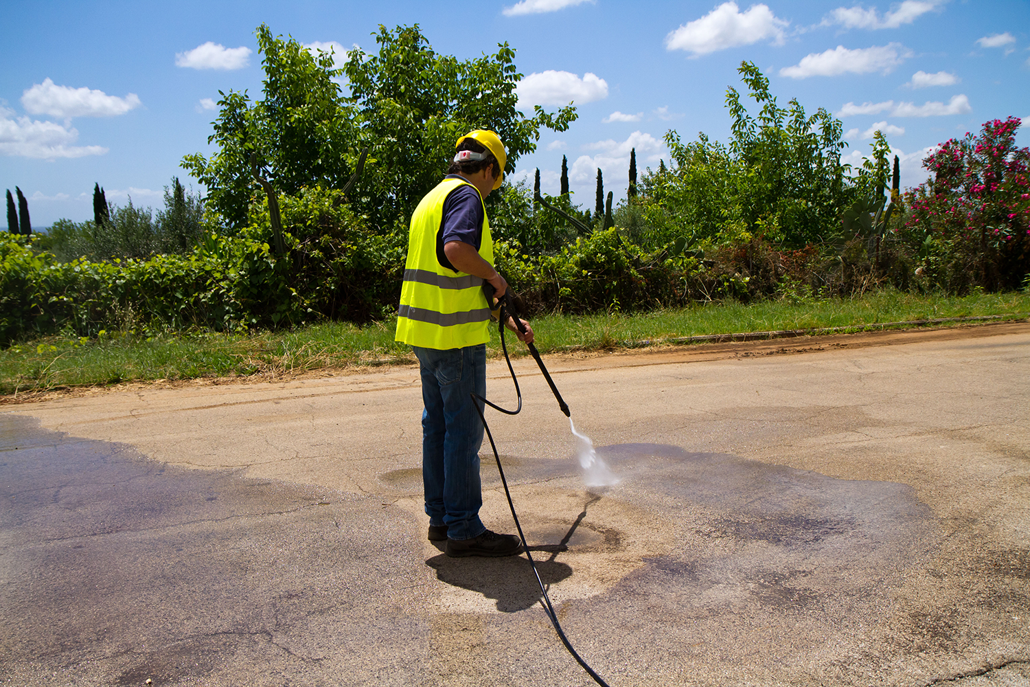 Worker in a yellow safety vest pressure-washing a road under a sunny sky.