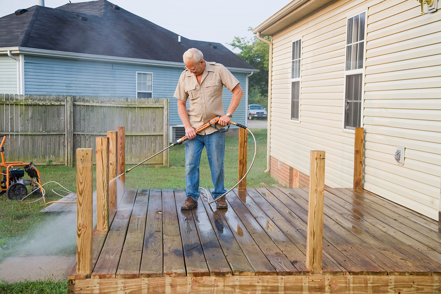 Man pressure-washing a wooden deck beside a beige house and blue yard fence