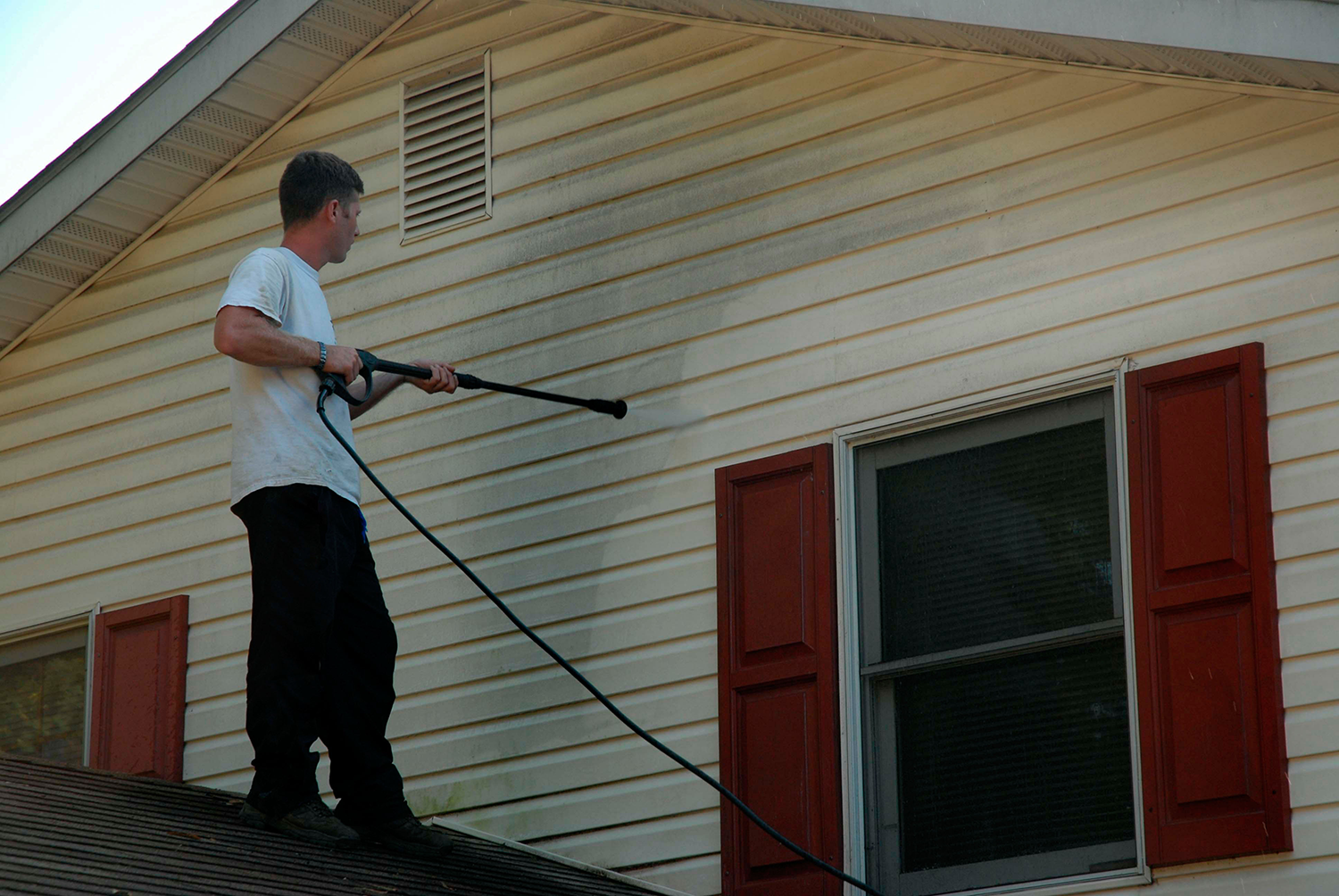 Person power-washing the side of a house with white siding and red shutters.