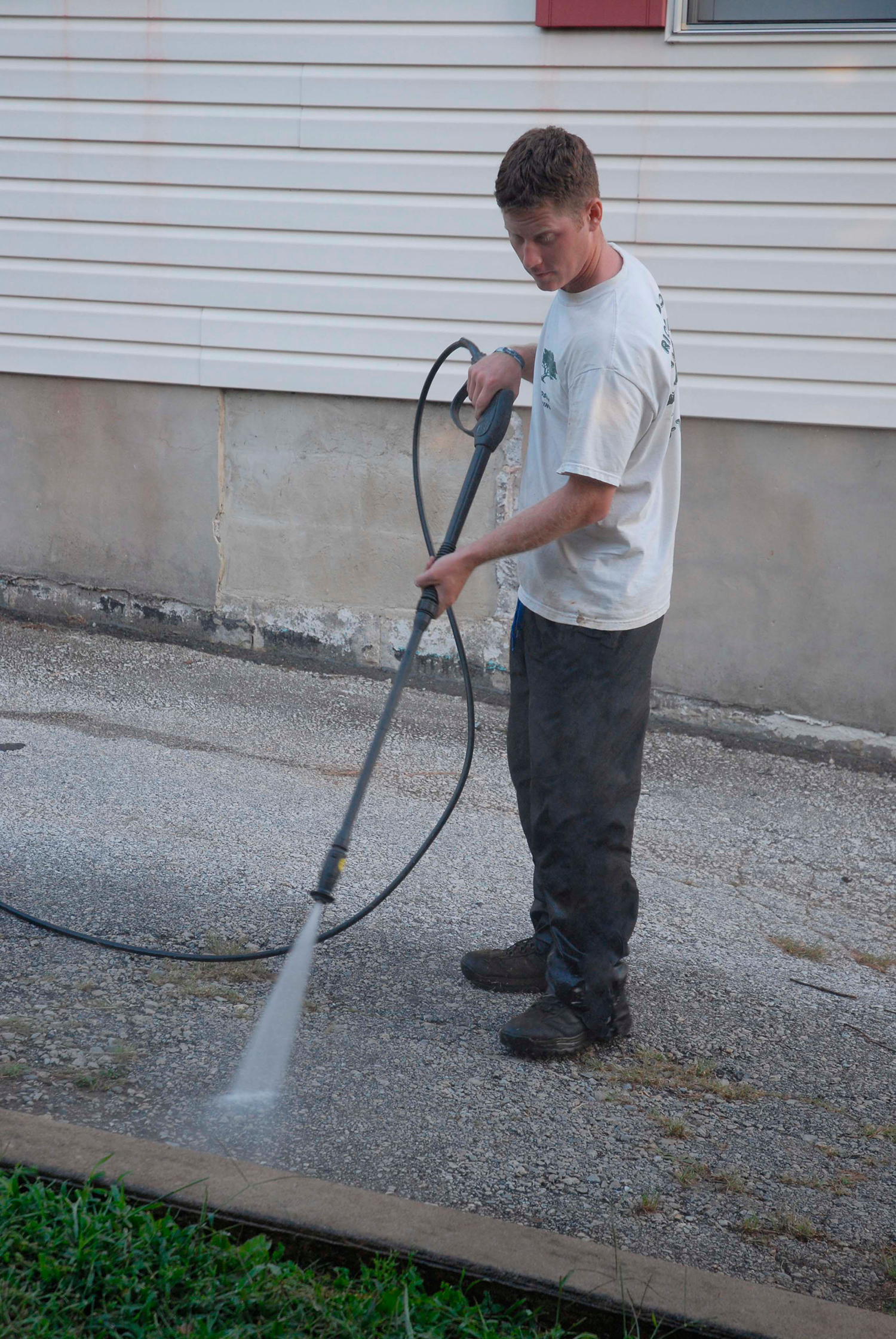 Person pressure-washing a gravel driveway beside a white house.