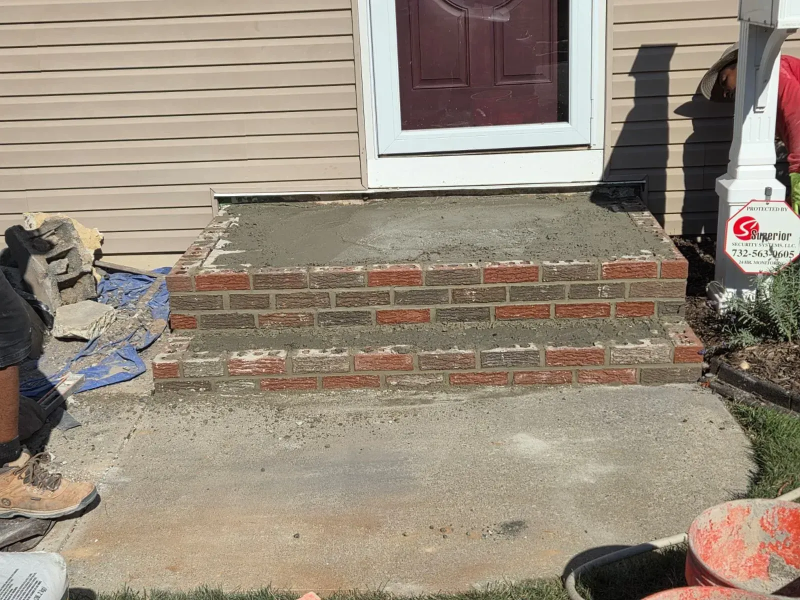 Brick front steps leading to a maroon door, with unfinished concrete patio and house siding