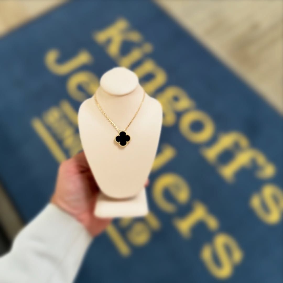 A hand holding a display with a black clover pendant necklace in front of a jeweler’s logo.