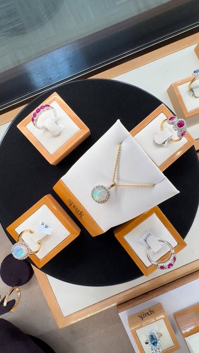 Jewelry display with rings and a necklace on a black circular platform. Gold and gemstone pieces in square display boxes.