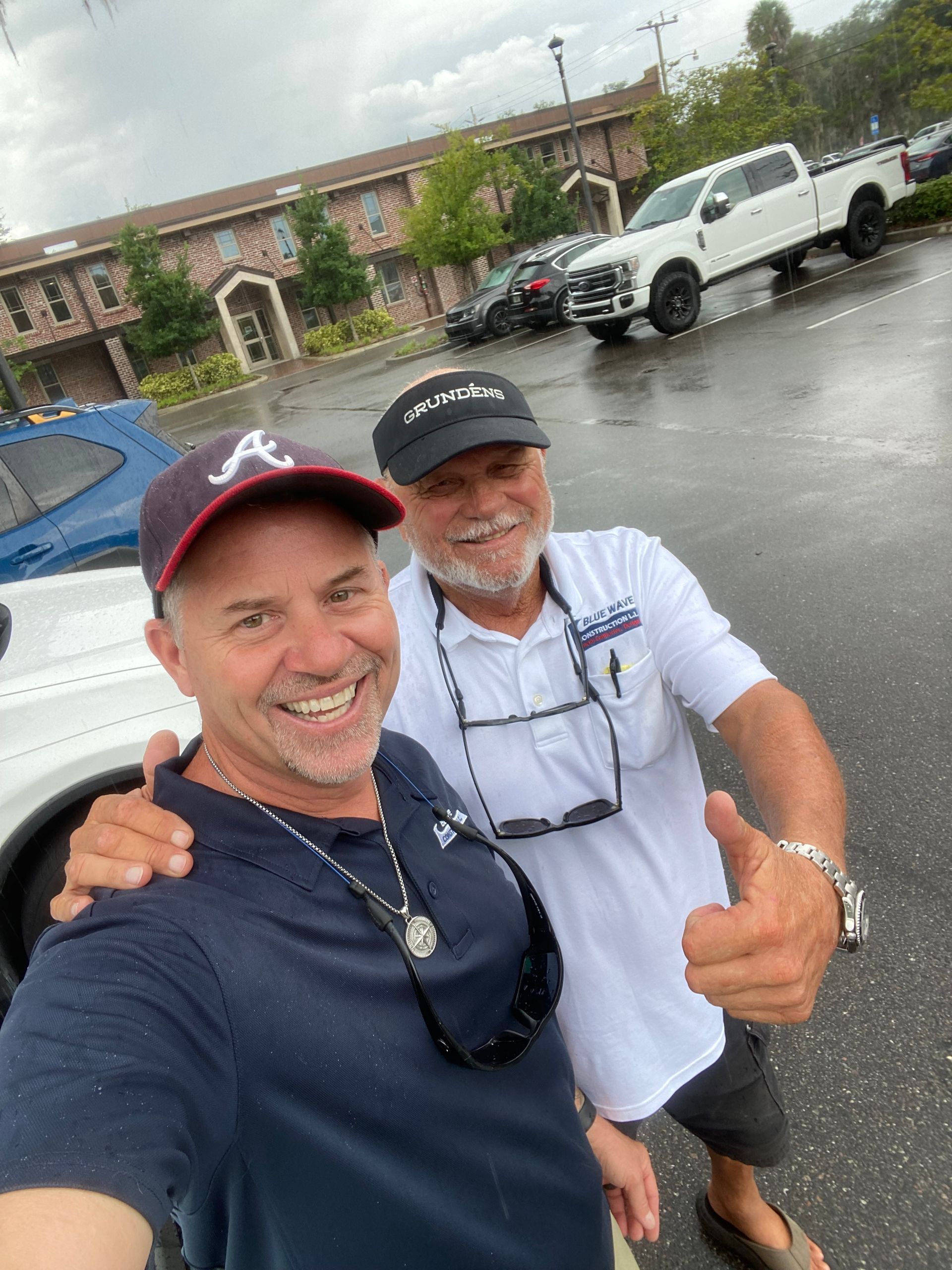 Two men smiling, one in an Atlanta Braves cap, taking a selfie outdoors with a truck and building in background.