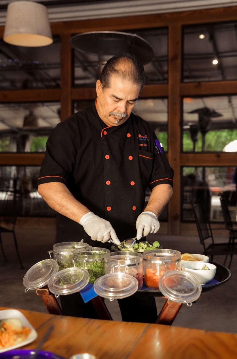 Person holding a cocktail garnished with citrus, surrounded by plates of food on a wooden table.