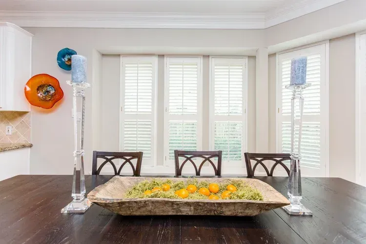 Dining room with dark table, floral centerpiece, three chairs, and white shuttered windows.