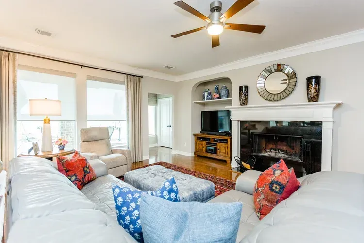 Bright living room with white sofas, blue pillows, fireplace, TV, wall clock, and ceiling fan