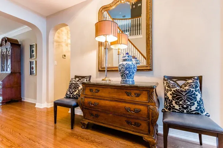Elegant entryway with wooden dresser, blue vase, lamps, mirror, and framed chair on hardwood floor