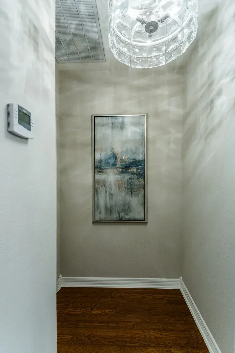 Narrow hallway with white walls, wood floor, ceiling light, and framed abstract artwork.