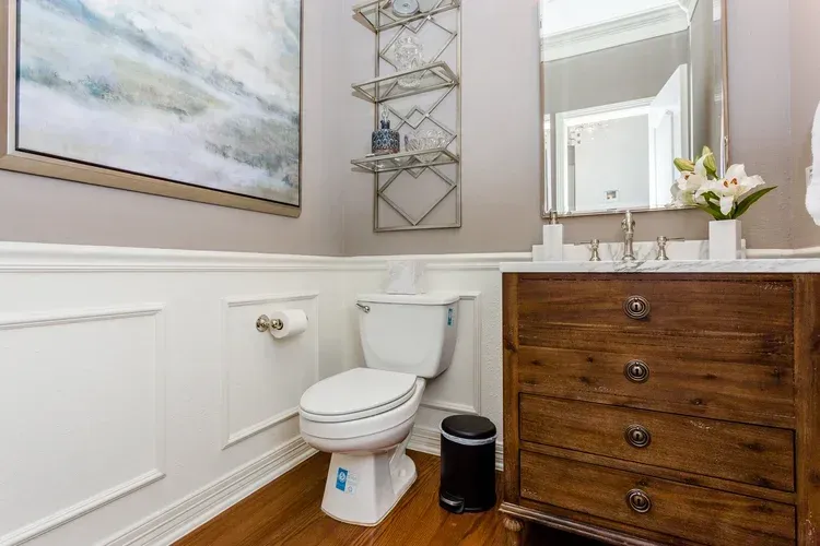 Small bathroom with white toilet, wood vanity, wall art, mirror, and flowers on the sink.