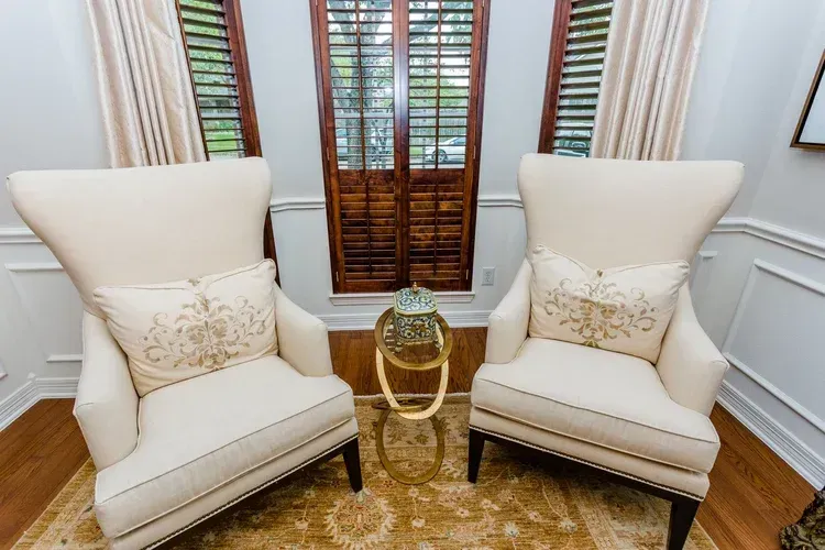 Two cream armchairs with pillows beside a small round table in a bright room with wooden shutters.
