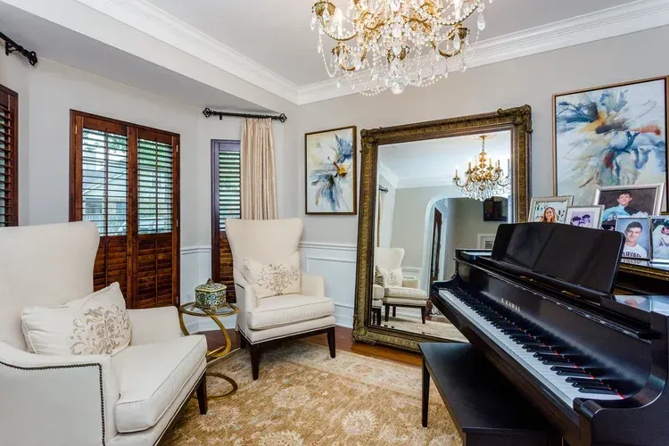 Elegant living room with a grand piano, gold-framed mirror, white chairs, and chandelier lighting