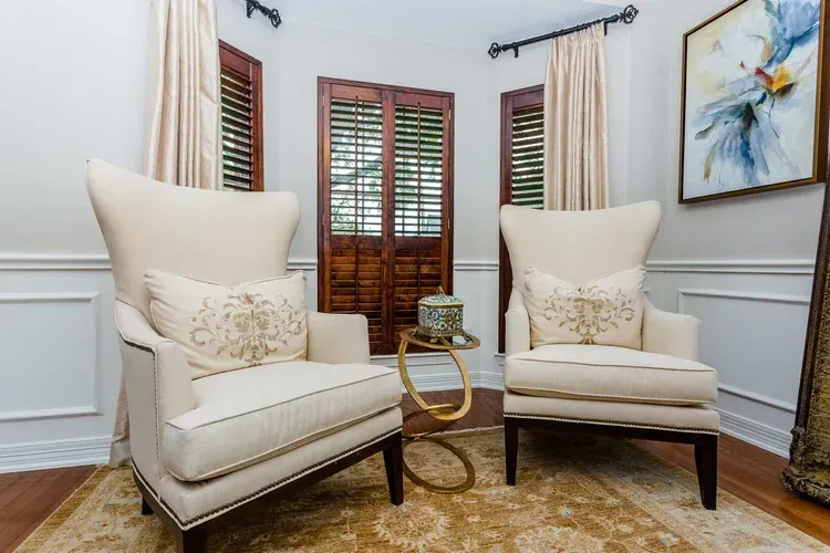 Two cream wingback chairs in a bright, elegant room with white walls, wood shutters, and a small side table.