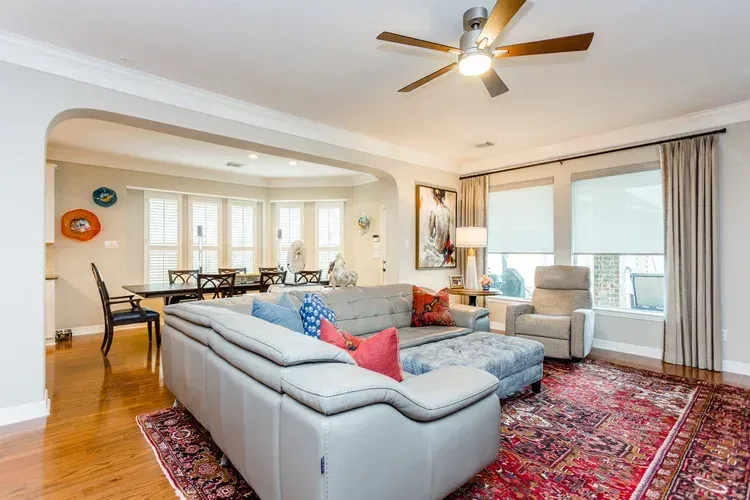 Bright living room with gray sectional, patterned rug, ceiling fan, and open dining area.