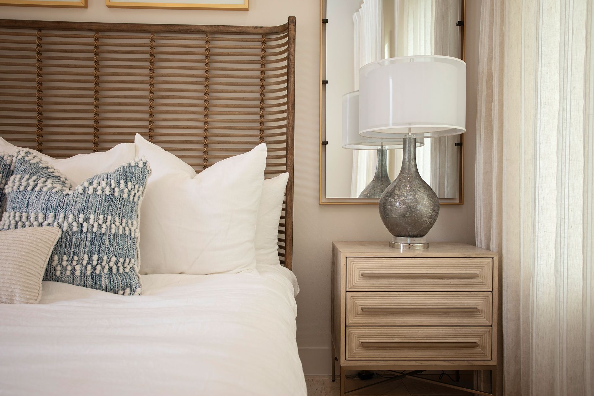 Cozy bedroom with white bedding, woven headboard, wooden nightstand, and silver lamp near sheer curtains