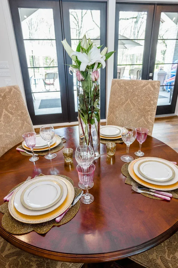 Elegant dining table set for four with gold-trimmed plates and a floral centerpiece by large windows
