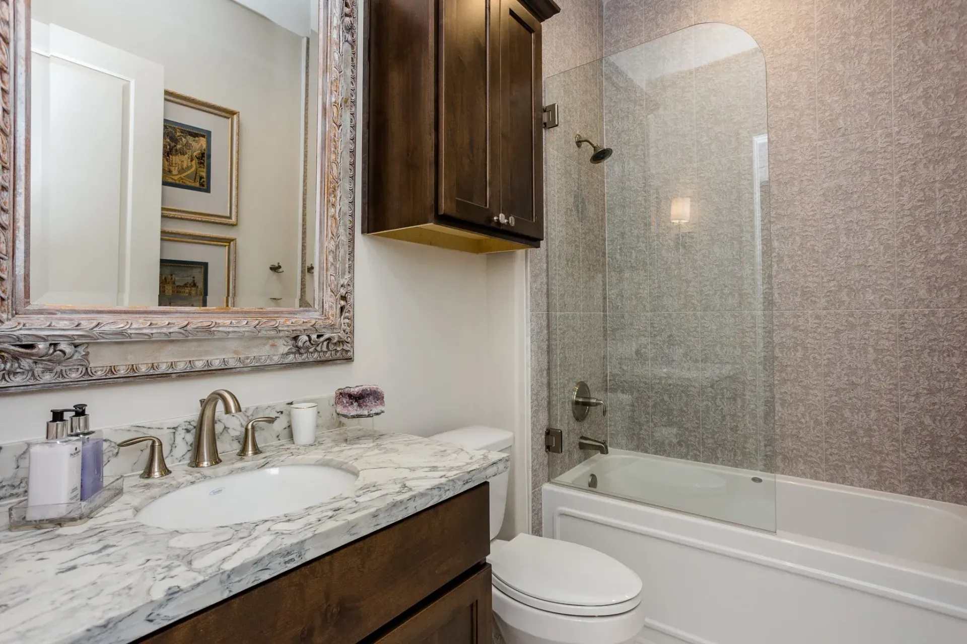 Modern bathroom with marble vanity, large mirror, wood cabinet, toilet, and bathtub with glass shower enclosure