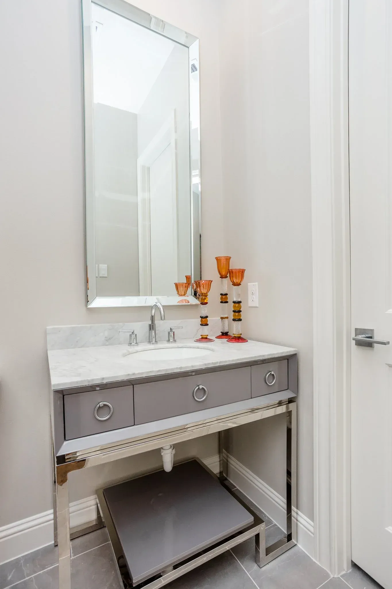 White bathroom vanity with a mirror, marble top, decorative bottles, and an open shelf with a folded rug.