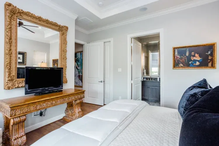 Bright bedroom with white bed, blue pillows, ornate gold mirror, and TV on a carved dresser