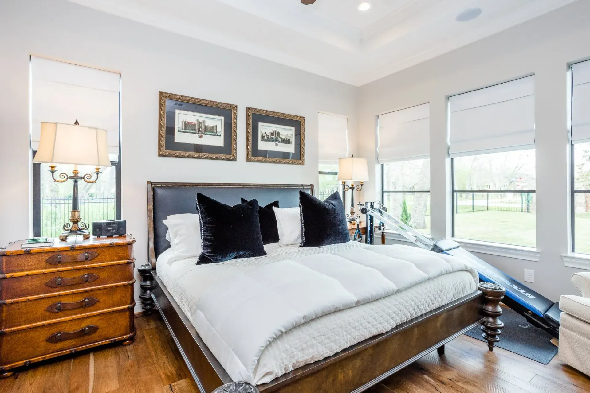 Bright bedroom with a large bed, wooden dresser, framed art, and tall windows with blinds.