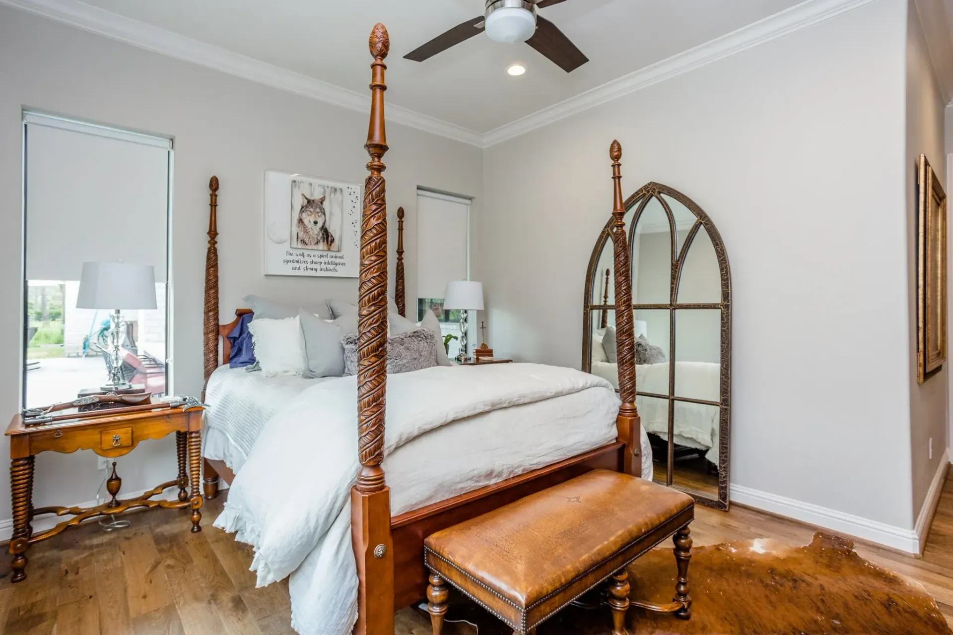 Bright bedroom with carved wood bed, bench, vanity, full-length mirror, and ceiling fan on gray walls.