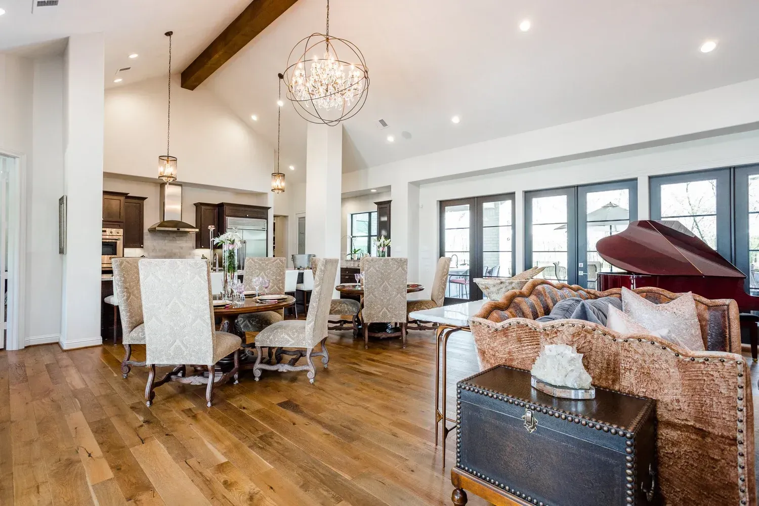 Bright open-concept living room and kitchen with hardwood floors, white chairs, and a patterned sofa under a chandelier