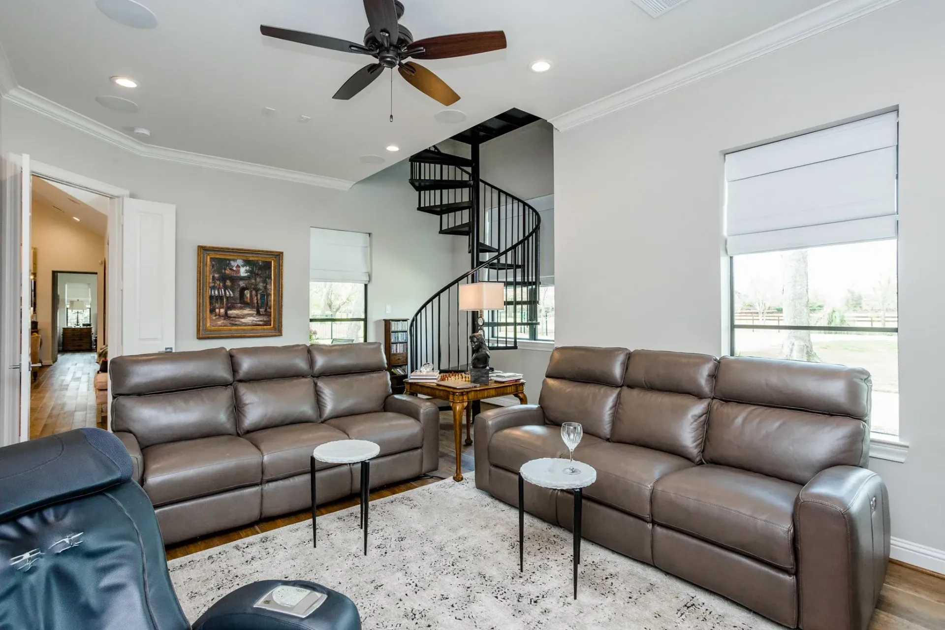 Living room with brown leather sofas, spiral staircase, ceiling fan, and bright windows