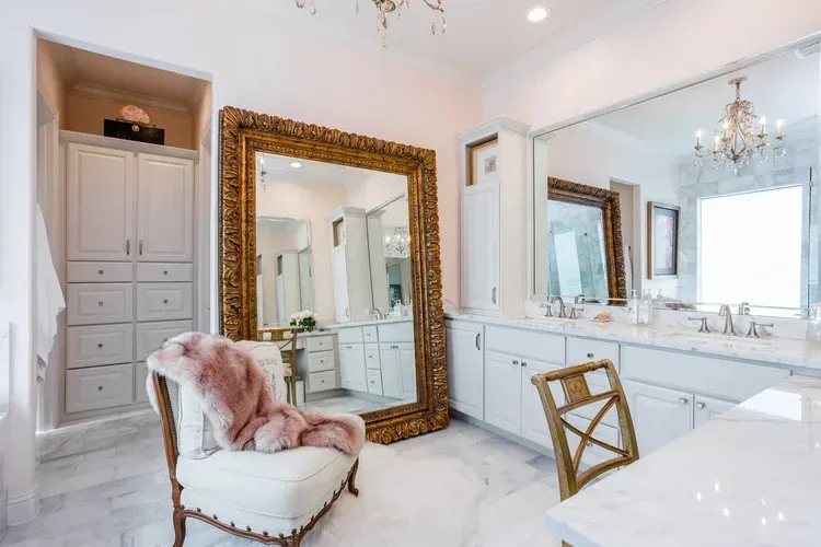Bright white luxury bathroom with a gold mirror, pink accent chair, and chandelier.