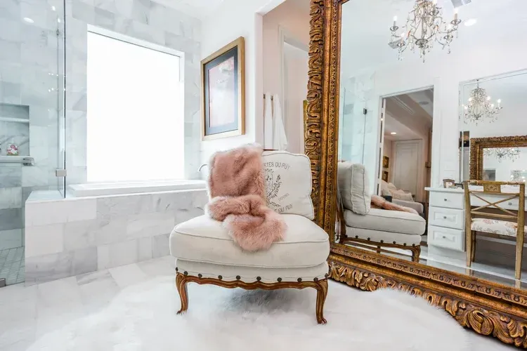 Bright white vanity room with ornate gold mirror, tufted chair, chandelier, and blush throw blanket
