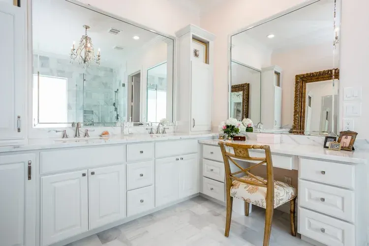Bright white vanity area with mirrored cabinets, marble counters, and a wicker chair in a luxurious bathroom.