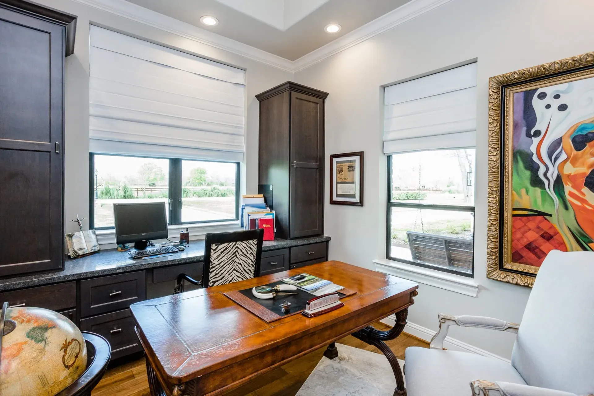 Bright home office with dark wood desk, computer, colorful art, and two windows with blinds