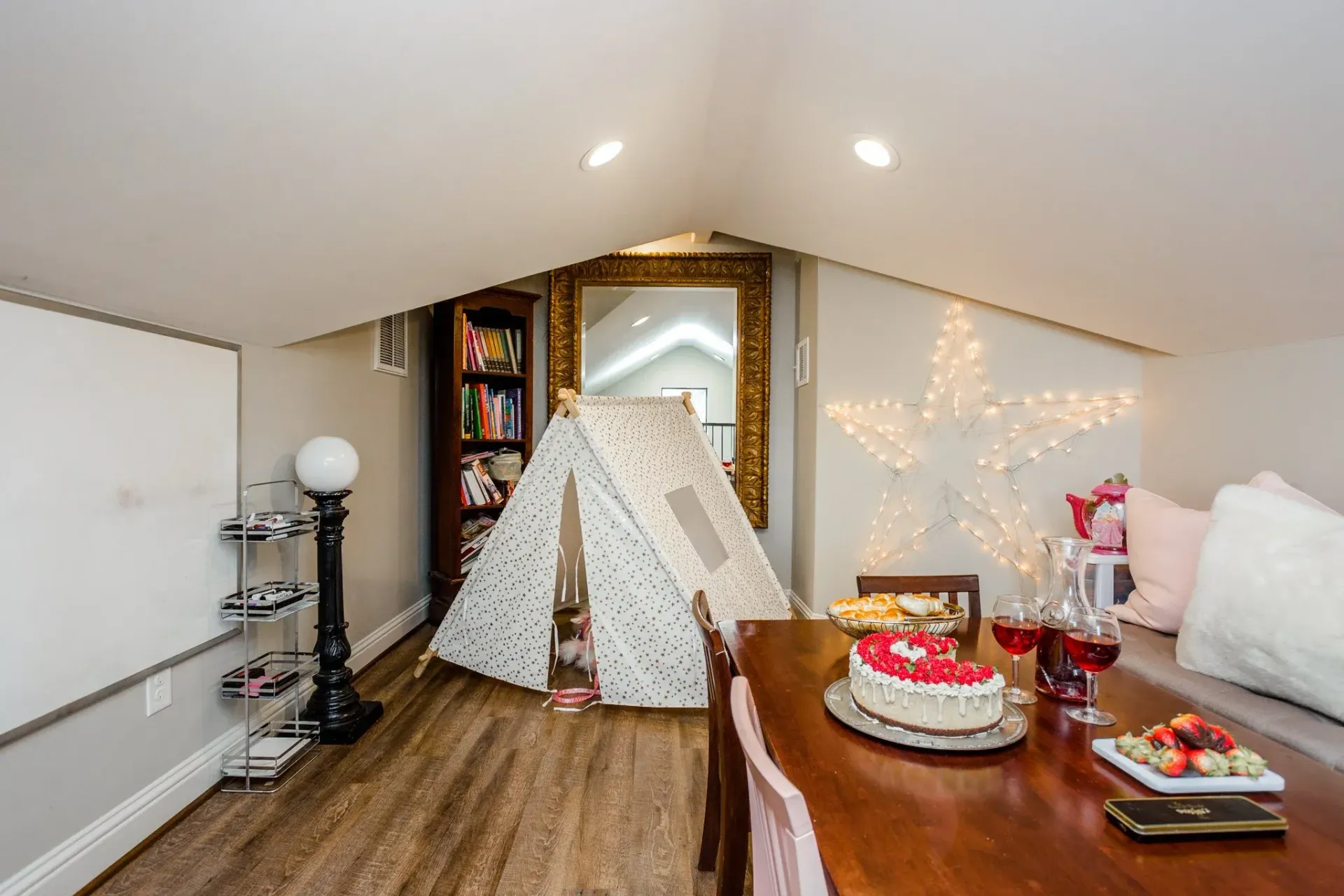 Attic room with a teepee, wooden table, and birthday cake on a sloped ceiling under warm lights