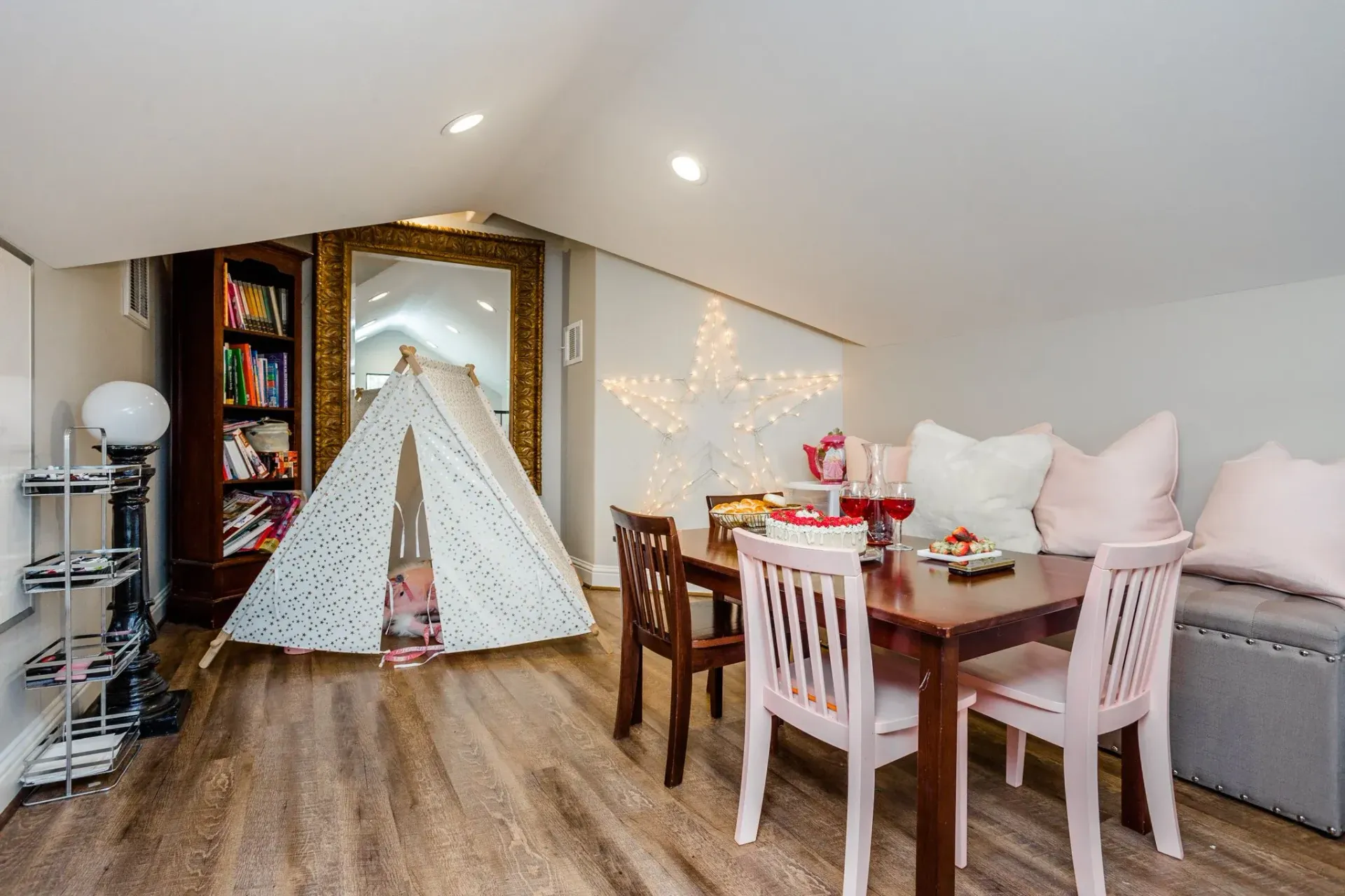 Cozy attic room with a teepee tent, dining table, sofa, and bookshelf under soft lighting