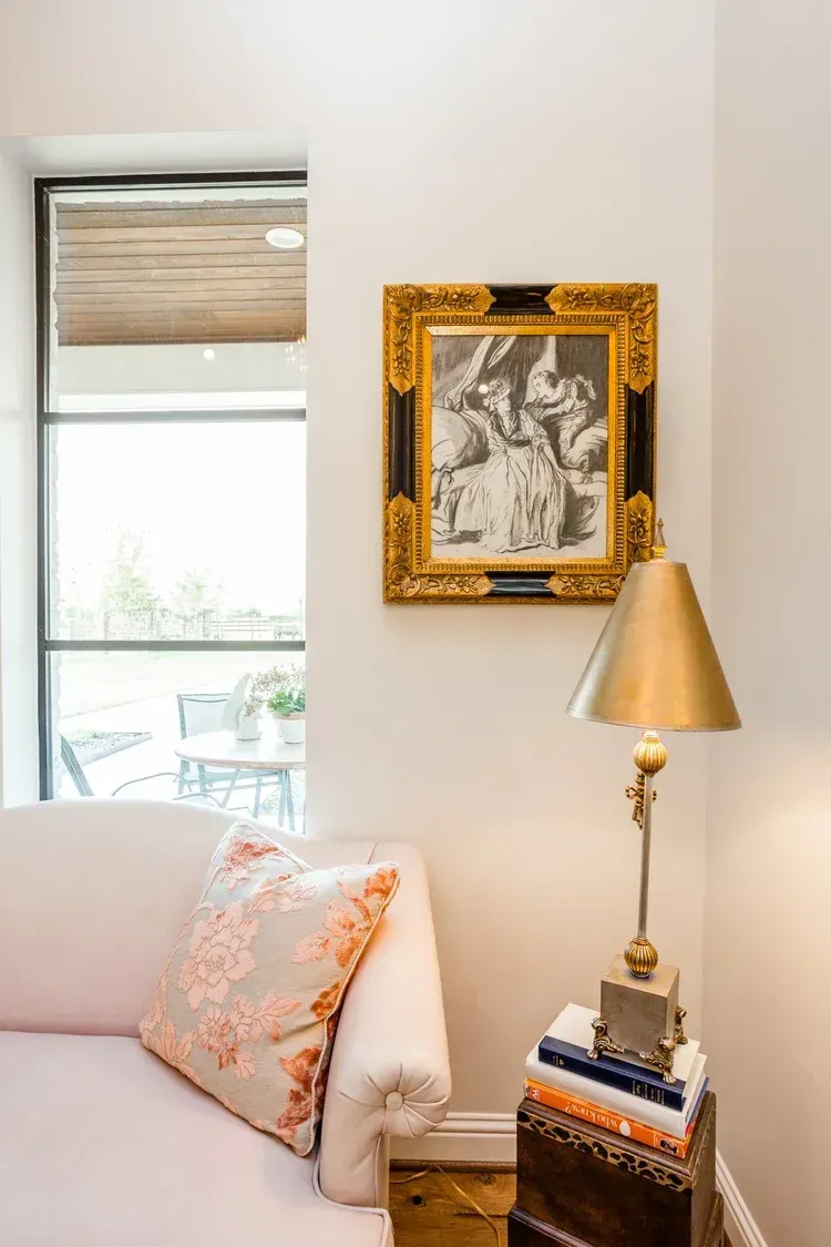 Elegant living room corner with gold-framed artwork, brass lamp, window, and a pale sofa with coral pillow