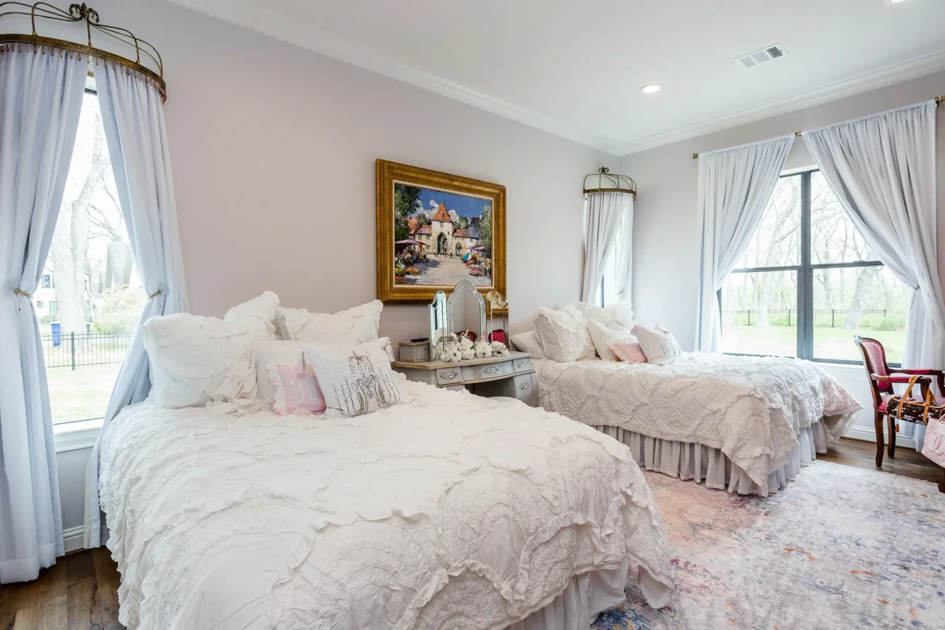 Bright white bedroom with two beds, sheer curtains, and a floral rug, lit by large windows.
