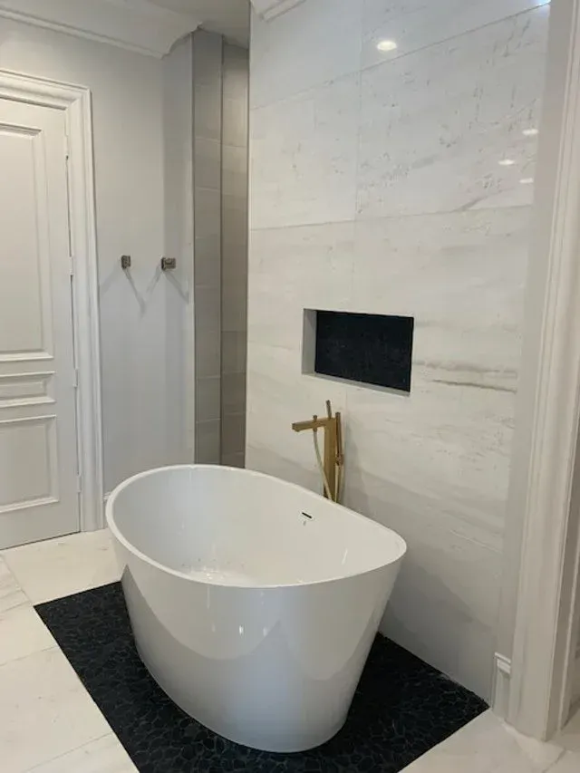 Modern bathroom with freestanding white tub, black rug, and gold faucet against marble wall.
