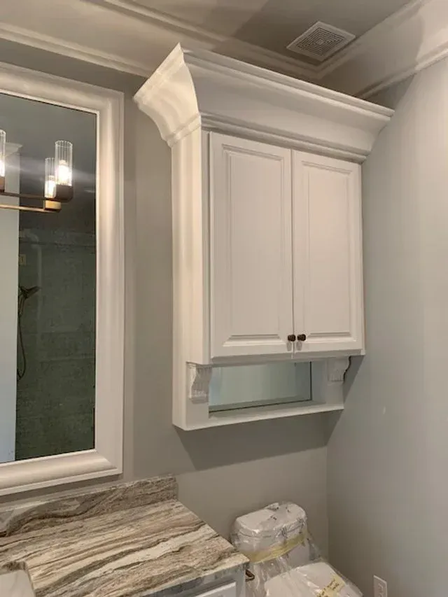 White bathroom cabinet above a marble countertop and window, with ornate crown molding.