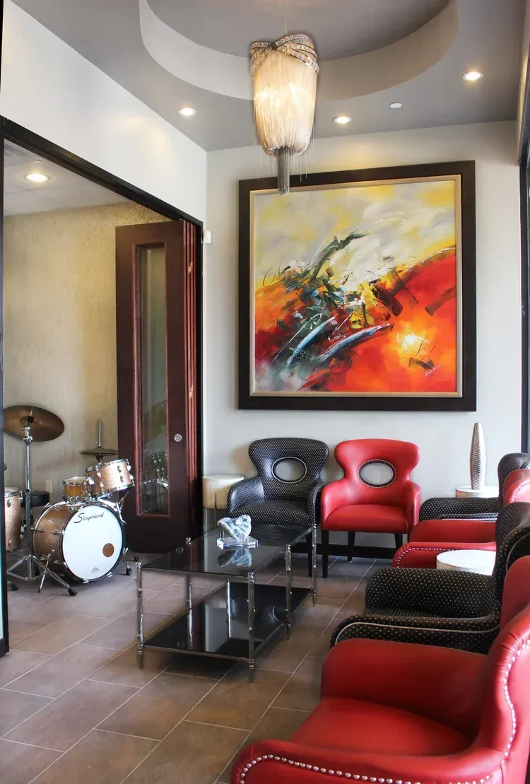 Modern lounge with red and black chairs, abstract wall art, glass table, and a drum set in the corner.