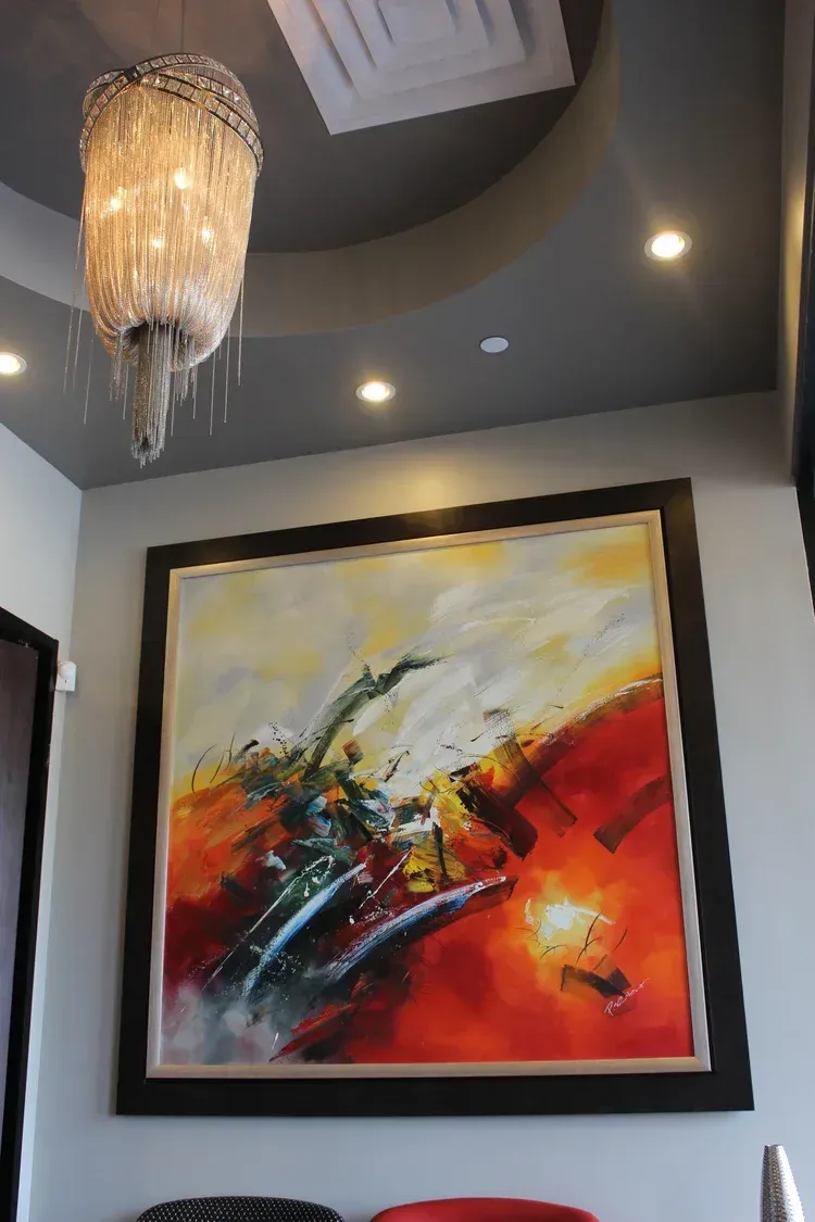 Framed abstract painting with red, orange, and yellow tones hanging on a gray wall under a chandelier