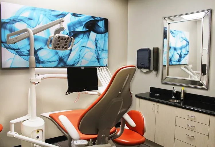 Modern dental exam room with orange chair, mounted monitor, sink, and blue wall art