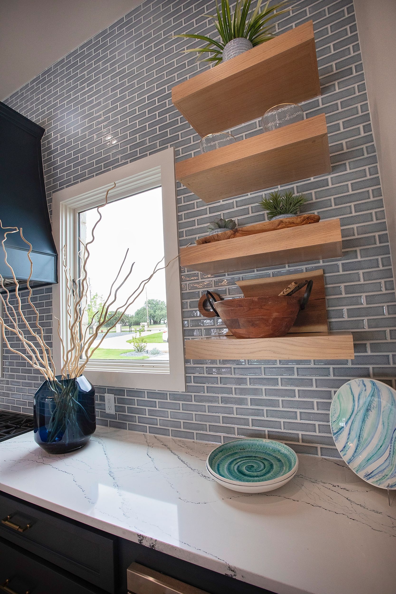 Modern kitchen wall with floating wood shelves, gray tile, and a white countertop by a window